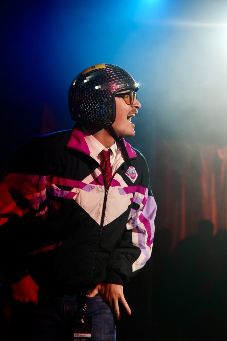Mann in bunter Sportjacke und Helm, lächelt enthusiastisch auf einer Bühne.