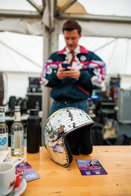 Ein glänzender Motorradhelm liegt auf einem Tisch, im Hintergrund ein Mann mit Smartphone.