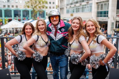 Eine Gruppe von Cheerleaderinnen posiert mit einem Mann in einem bunten Outfit und Sonnenbrille.