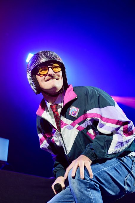 Mann in Retro-Jacke und Helm performt auf einer Bühne mit bunten Lichtern.